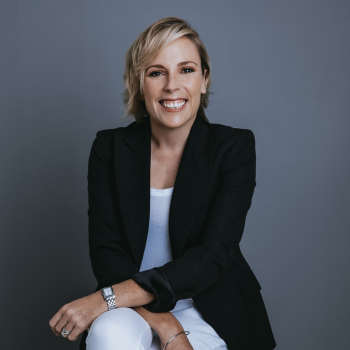 Smiling woman with short blonde hair, wearing a black blazer and white outfit, seated against a plain grey backgroud.